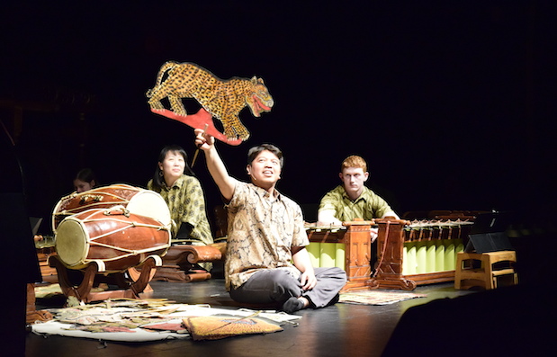 A puppeteer holds a tiger puppet while instrumentalists play behind him.