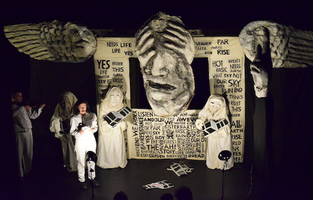 A large black and white puppet with wings takes up the stage while a chorus of actors stand on stage.