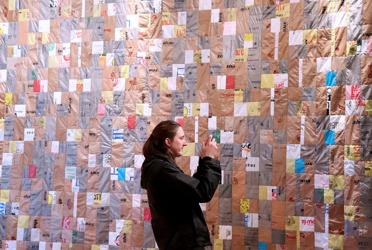 Someone stands before a large quilt made of plastic bags.